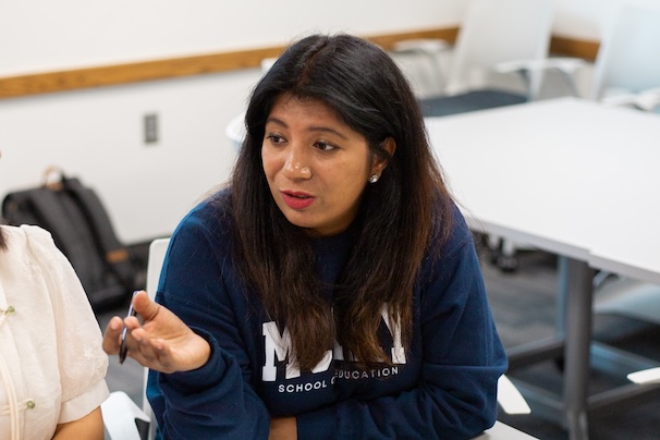 A woman wearing a blue sweatshirt with "School of Education" written on it is engaged in a discussion in a classroom setting. She is gesturing with her hand while speaking, with a thoughtful expression on her face. The background shows classroom furniture, including chairs and tables.